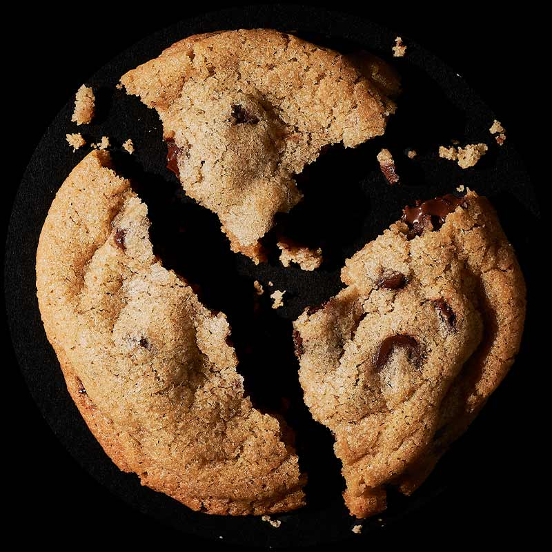 A chocolate chip cookie viewed from above, broken into three unequal pieces, surrounded by scattered crumbs.